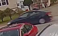 Two cars on a suburban street: a red car in the foreground with a dark sedan behind, houses and trees in the background.