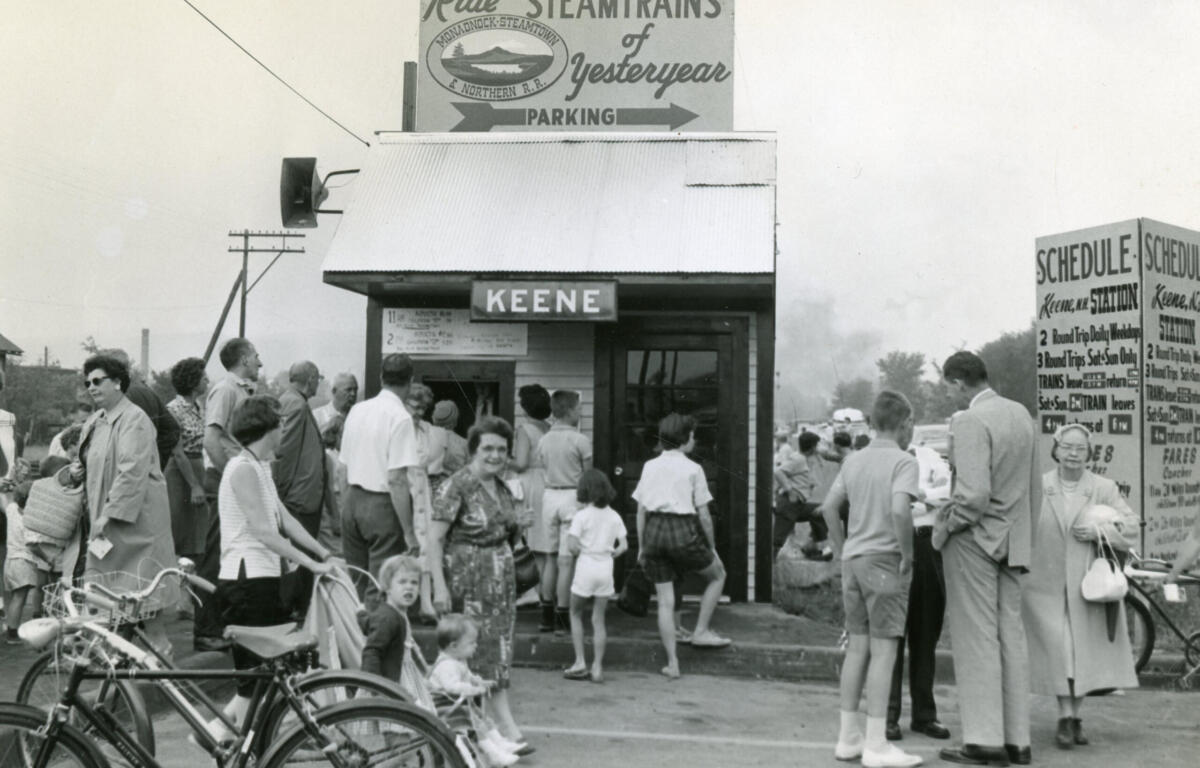 Crowd gathers around a small ticket or kiosk booth labeled KEENE at a vintage street fair; bicycles parked in foreground.