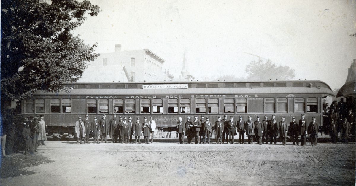 Vintage passenger railcar with a long row of windows; a line of men in early 20th-century attire stand along a dirt platform in front of the car.
