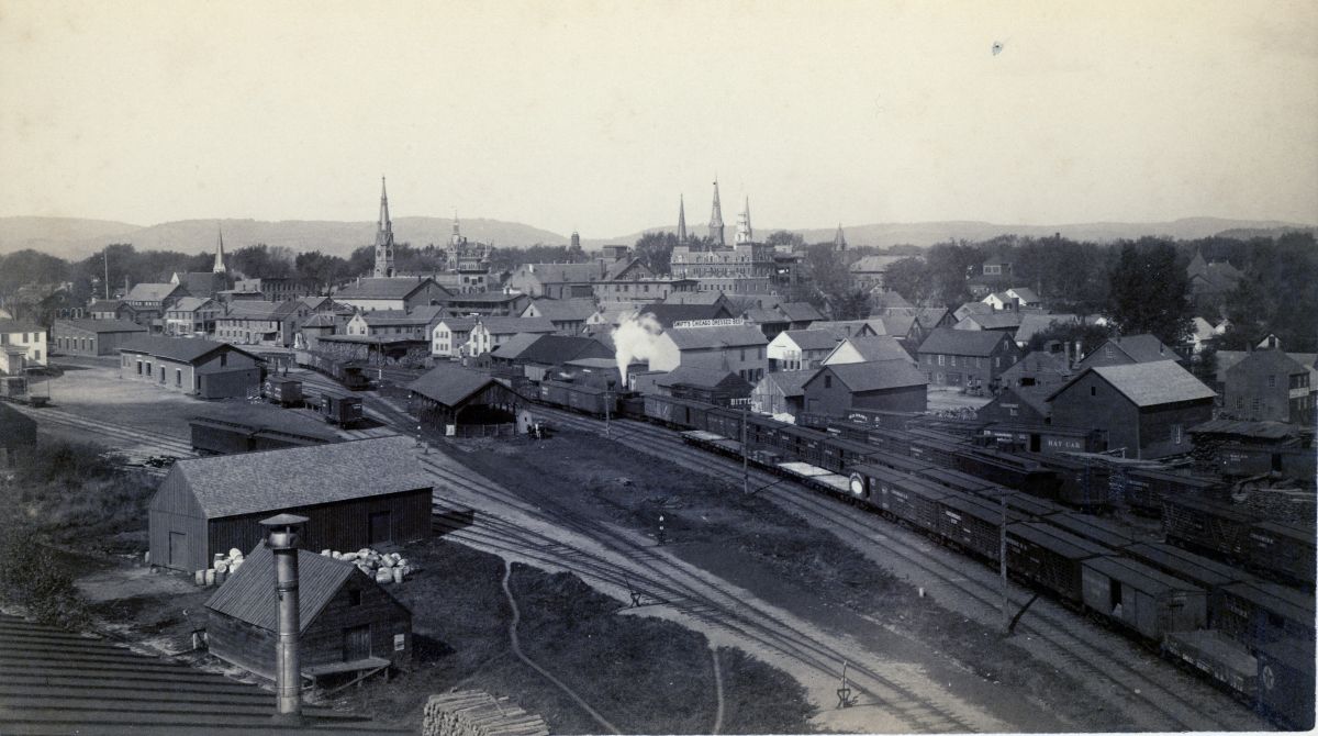 Historic railway yard in a small town with steam locomotive, tracks, warehouses, and church spires in the background.