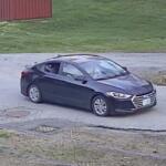 Dark gray four-door sedan driving on a wet road, driver visible through the window; grassy area and a red shed in the background.