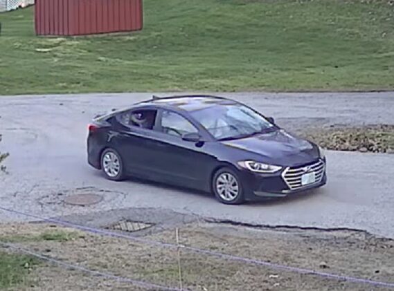 Dark gray four-door sedan driving on a wet road, driver visible through the window; grassy area and a red shed in the background.