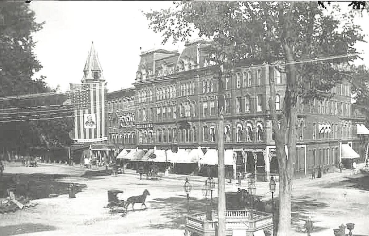 Historic city street with brick row houses, awnings along ground floors, a horse-drawn carriage in the foreground, and a tall church steeple in the background.