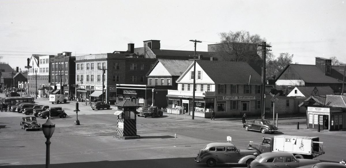 Black-and-white street scene of a mid-20th-century town with vintage cars and storefronts at a busy intersection, Crystal Restaurant visible. It shows a small downtown with a newsstand and pedestrians.