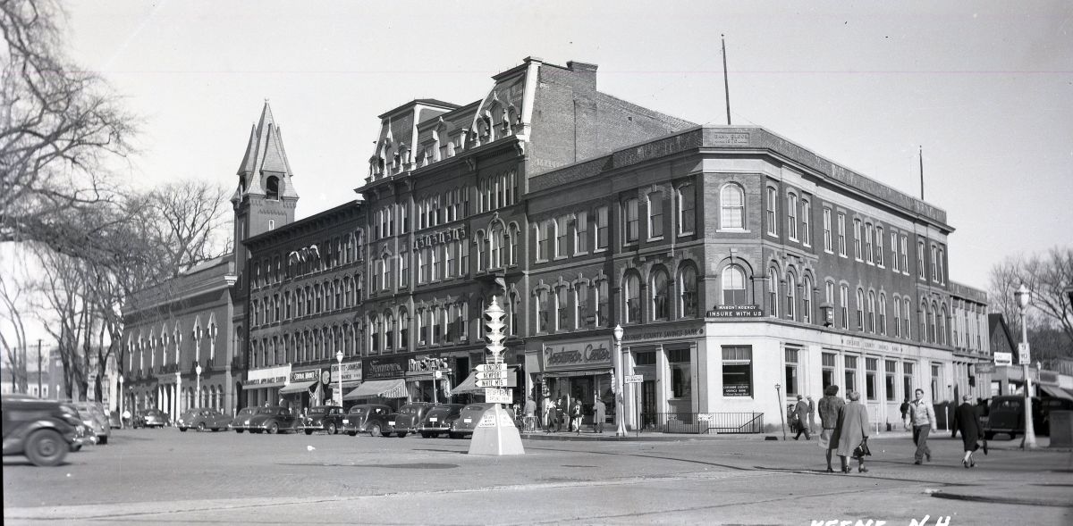 Row of brick 3- to 4-story commercial buildings with arched windows on a busy street; vintage cars, pedestrians, and a monument in the center.