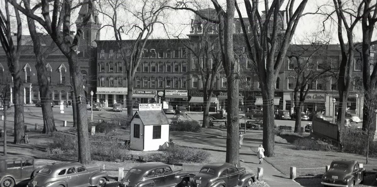 Black-and-white street scene with a small white kiosk in a park, bare trees, vintage cars, and storefronts in the background.
