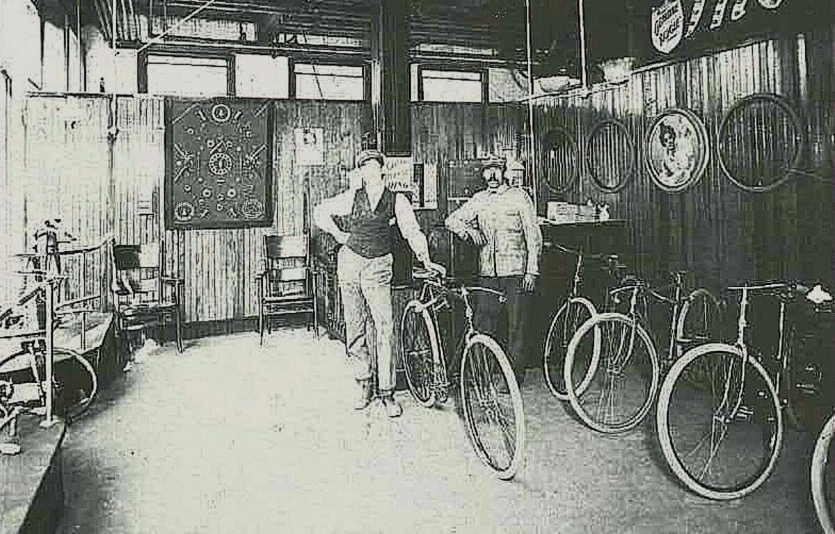 Two people stand with vintage bicycles inside a retro bike shop, with bikes lined along the right and wooden walls behind them.
