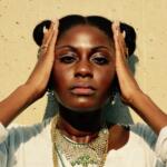 Close-up of a woman with hands framing her face, wearing multiple gold and green necklaces and earrings, against a light wall.