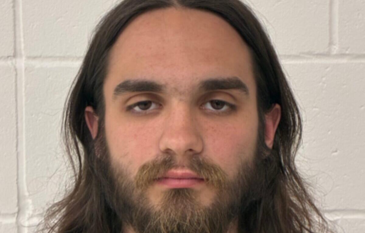 Mugshot-style portrait of a man with shoulder-length brown hair and a full beard, looking at the camera against a light gray concrete block wall behind him.