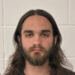 Mugshot-style portrait of a man with shoulder-length brown hair and a full beard, looking at the camera against a light gray concrete block wall behind him.
