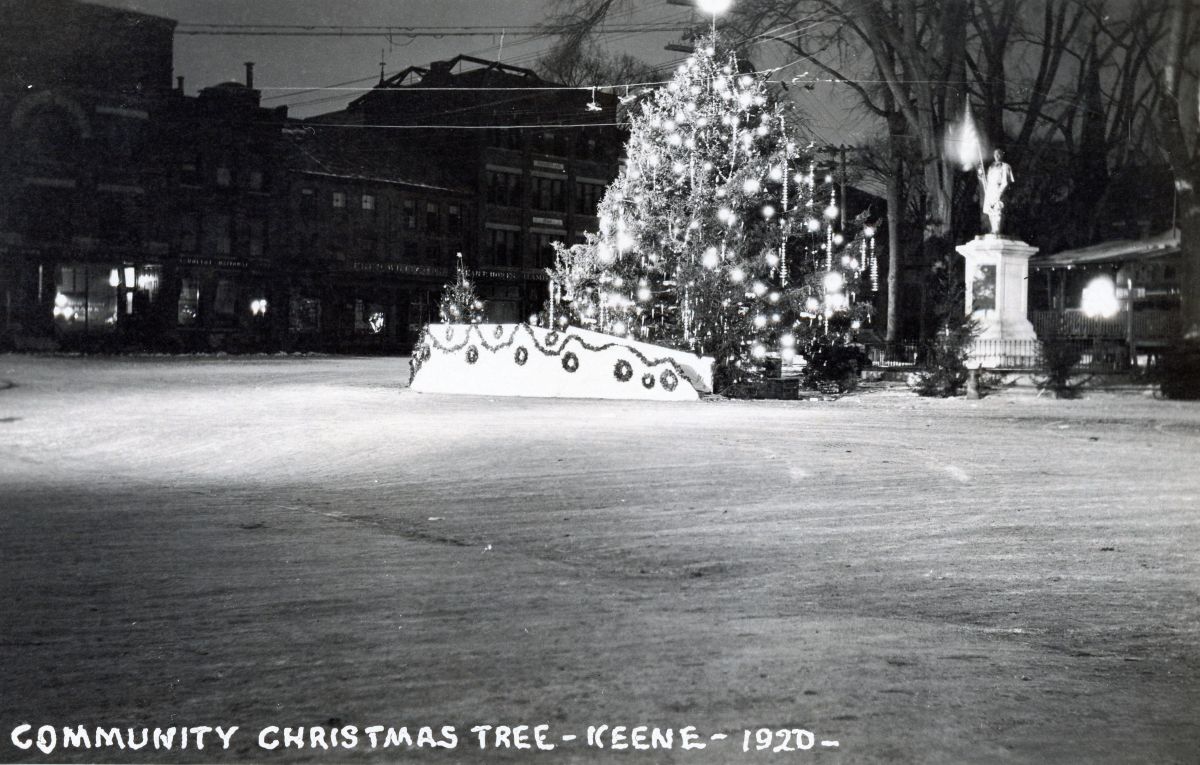 Night scene of a large illuminated Christmas tree in a town square, with garlands and wreaths, a statue to the right, and a snow-covered ground. Caption reads: 'Community Christmas Tree – Keene – 1920'.