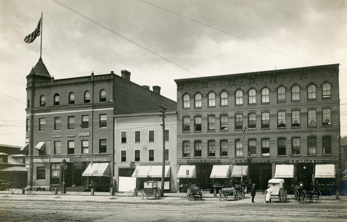 Row of three brick storefronts with striped awnings and horse-drawn carriages on a dirt street; American flag on the left building.