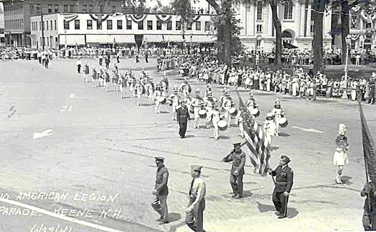 Black-and-white parade scene with soldiers marching and flag bearers along a city street, spectators lining the sidewalks.