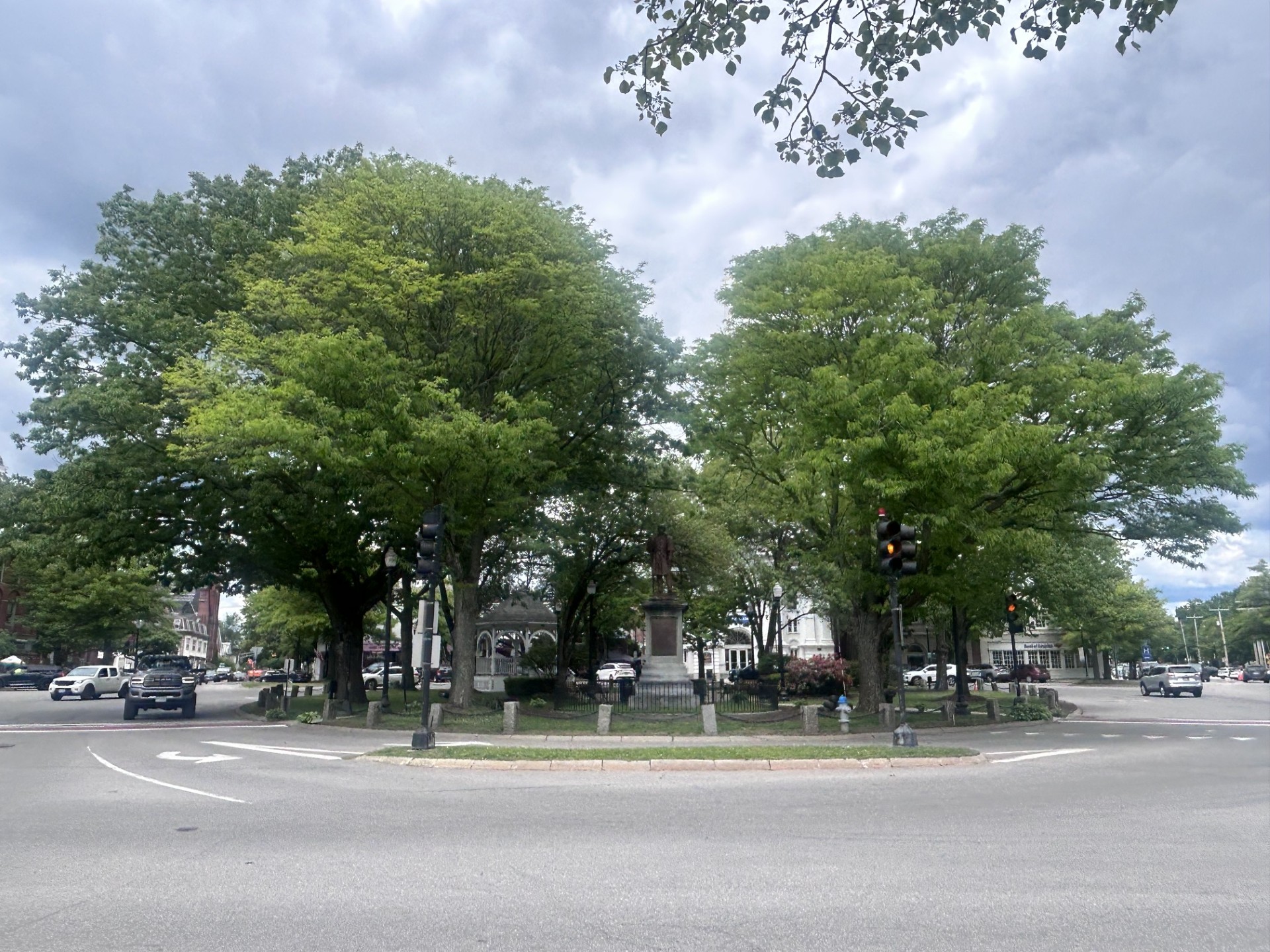 Green roundabout with a statue at the center, framed by large leafy trees and city streets around it to the sides.