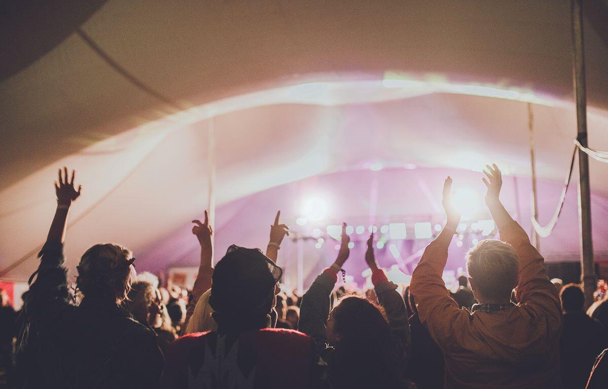 Crowd at a music festival inside a tent, hands raised toward a brightly lit stage with pink-purple lights.