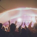 Crowd at a music festival inside a tent, hands raised toward a brightly lit stage with pink-purple lights.