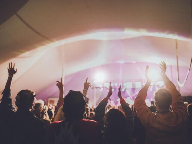 Crowd at a music festival inside a tent, hands raised toward a brightly lit stage with pink-purple lights.