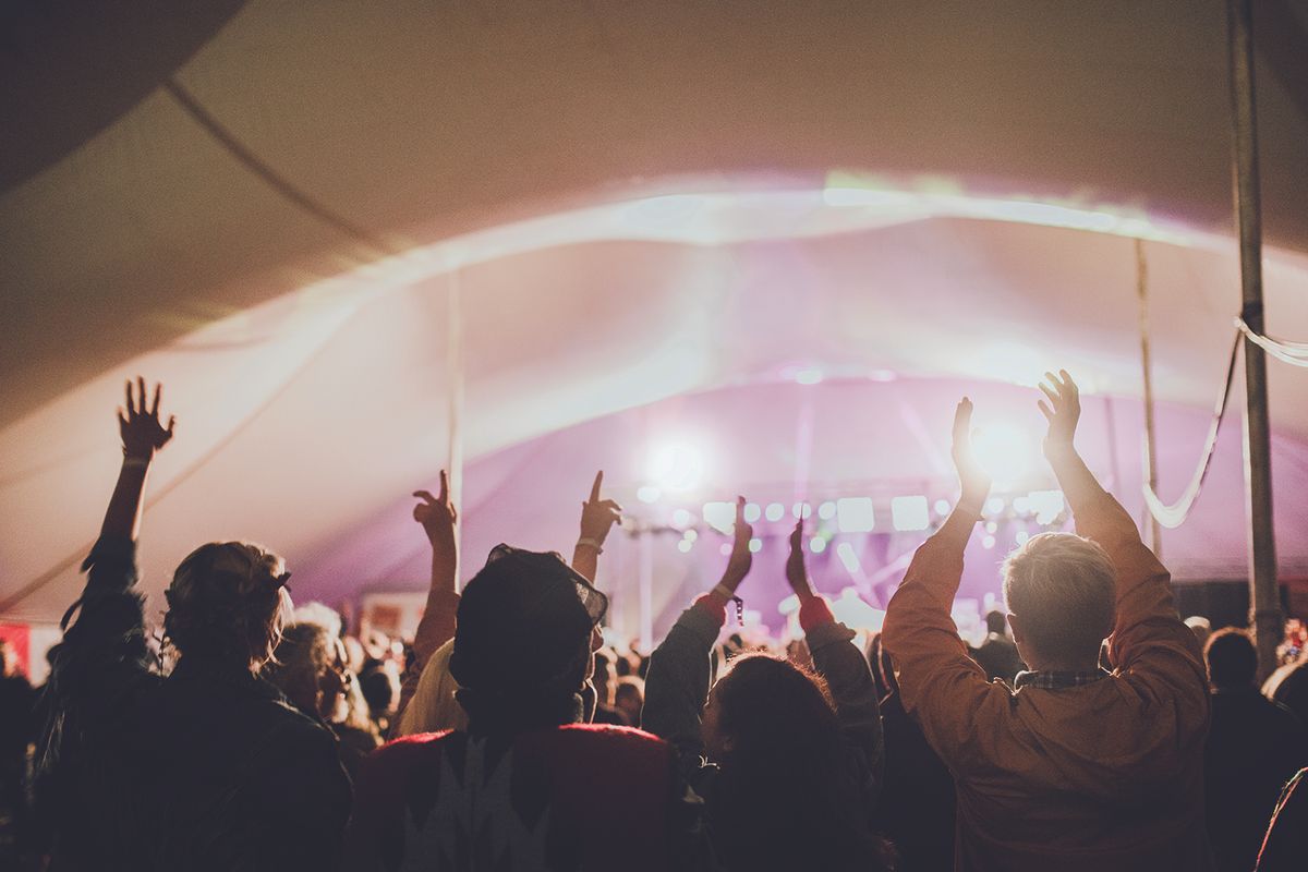 Crowd at a music festival inside a tent, hands raised toward a brightly lit stage with pink-purple lights.