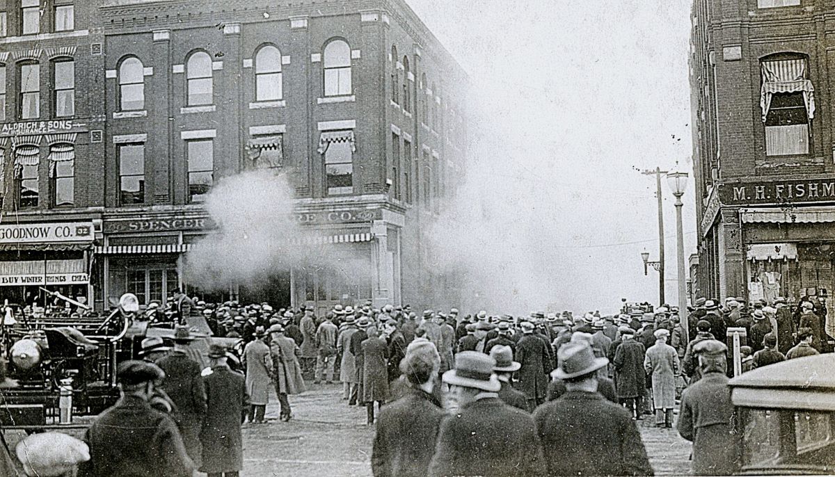 Crowd of men and women in 1920s attire on a brick‑building street as a smoke plume rises from a storefront in the center of the scene.