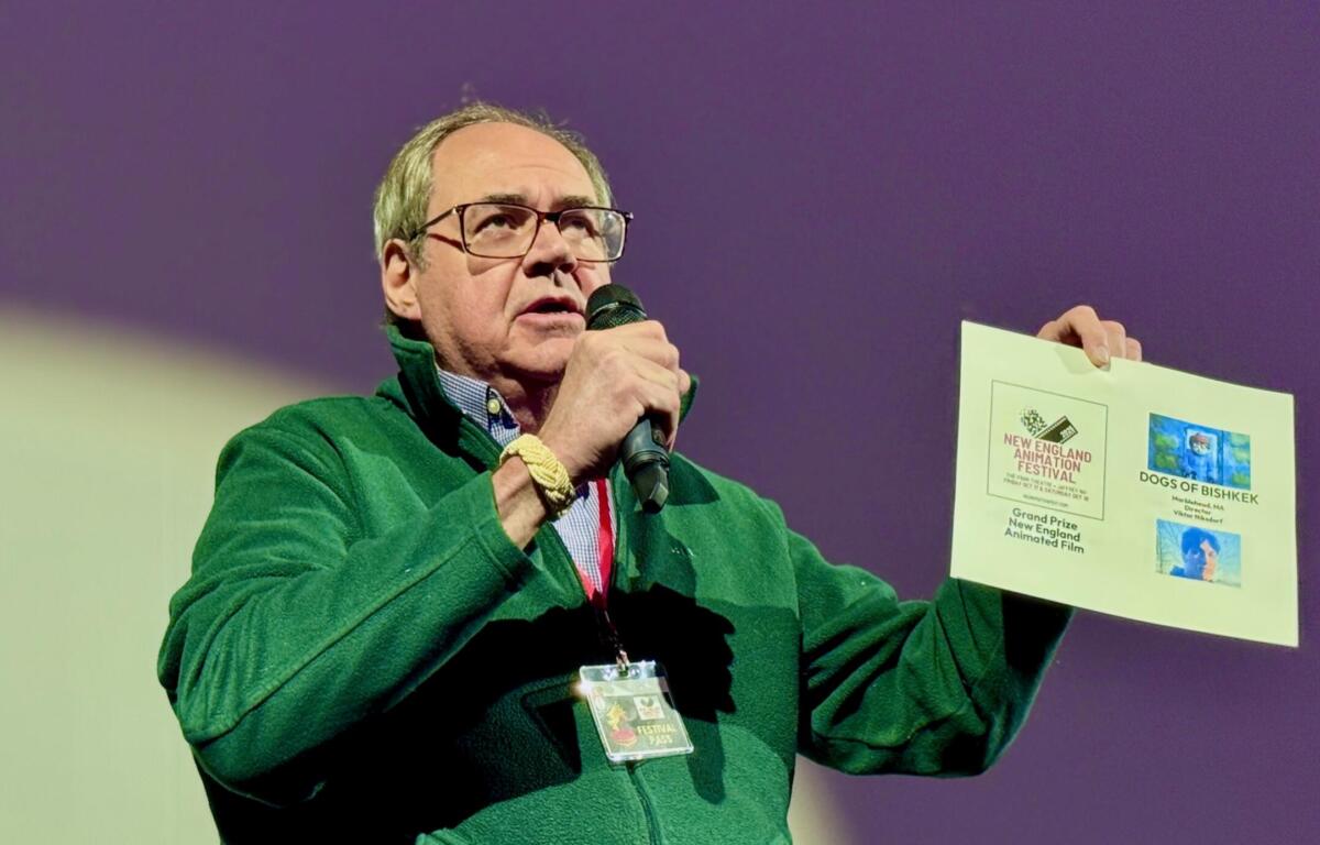 Man in a green jacket speaks into a microphone while holding a poster board with event flyers at a presentation venue.