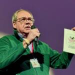 Man in a green jacket speaks into a microphone while holding a poster board with event flyers at a presentation venue.
