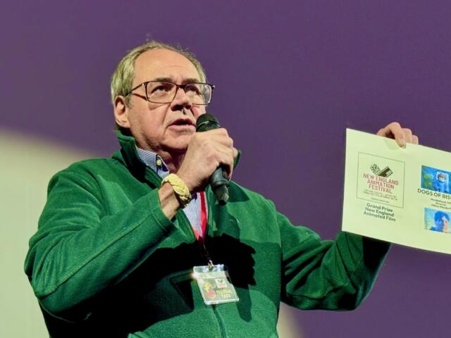 Man in a green jacket speaks into a microphone while holding a poster board with event flyers at a presentation venue.
