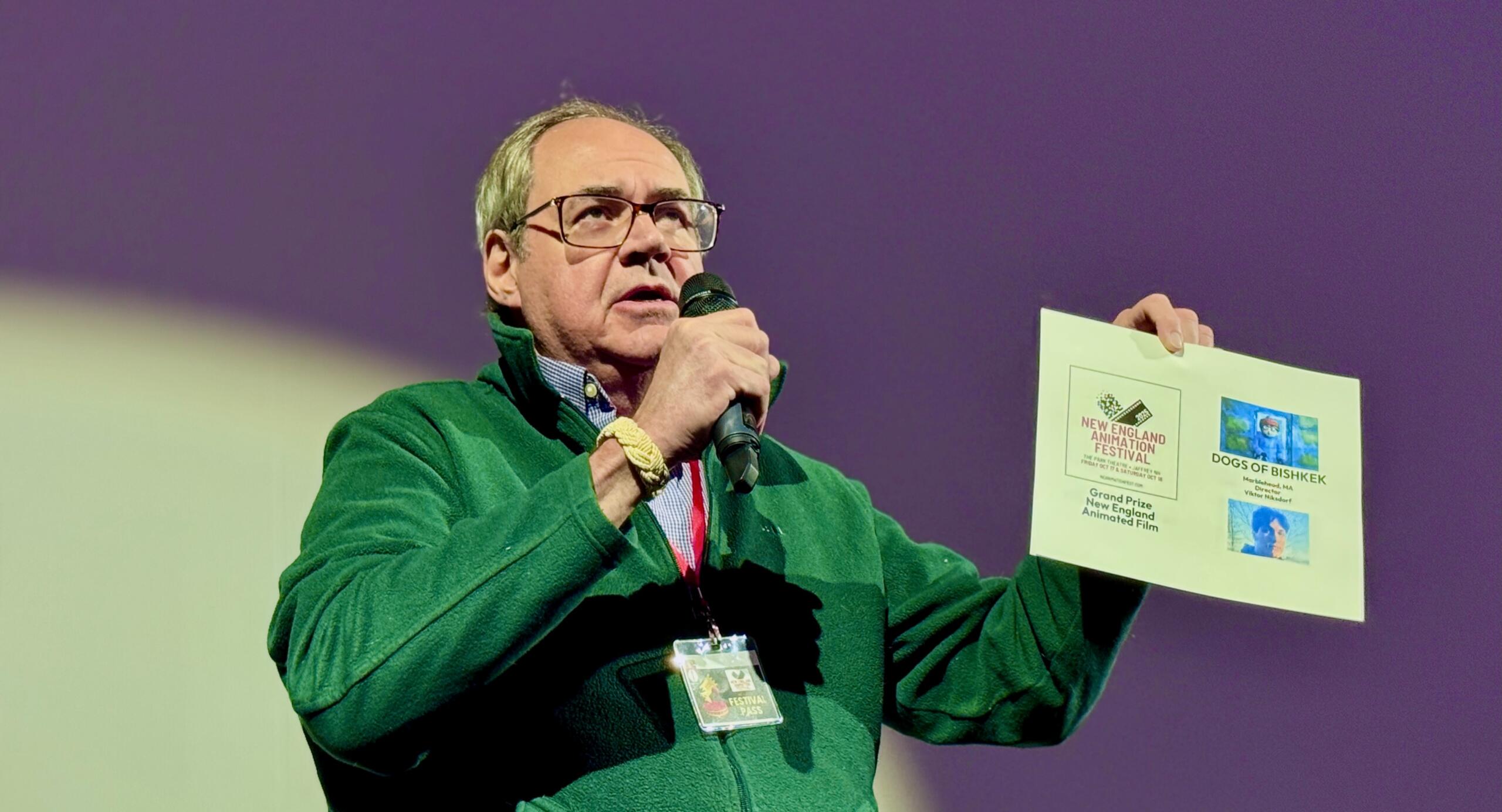 Man in a green jacket speaks into a microphone while holding a poster board with event flyers at a presentation venue.