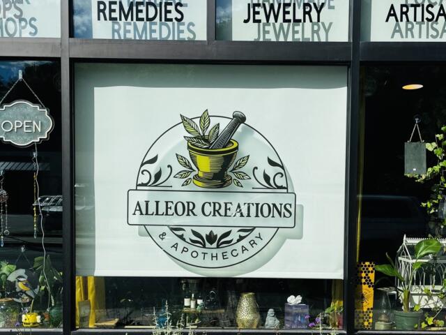 Storefront window display for Alleor Creations Apothecary featuring a circular logo with a mortar and pestle, leaves, and the text 'ALLEOR CREATIONS' and 'APOTHECARY'