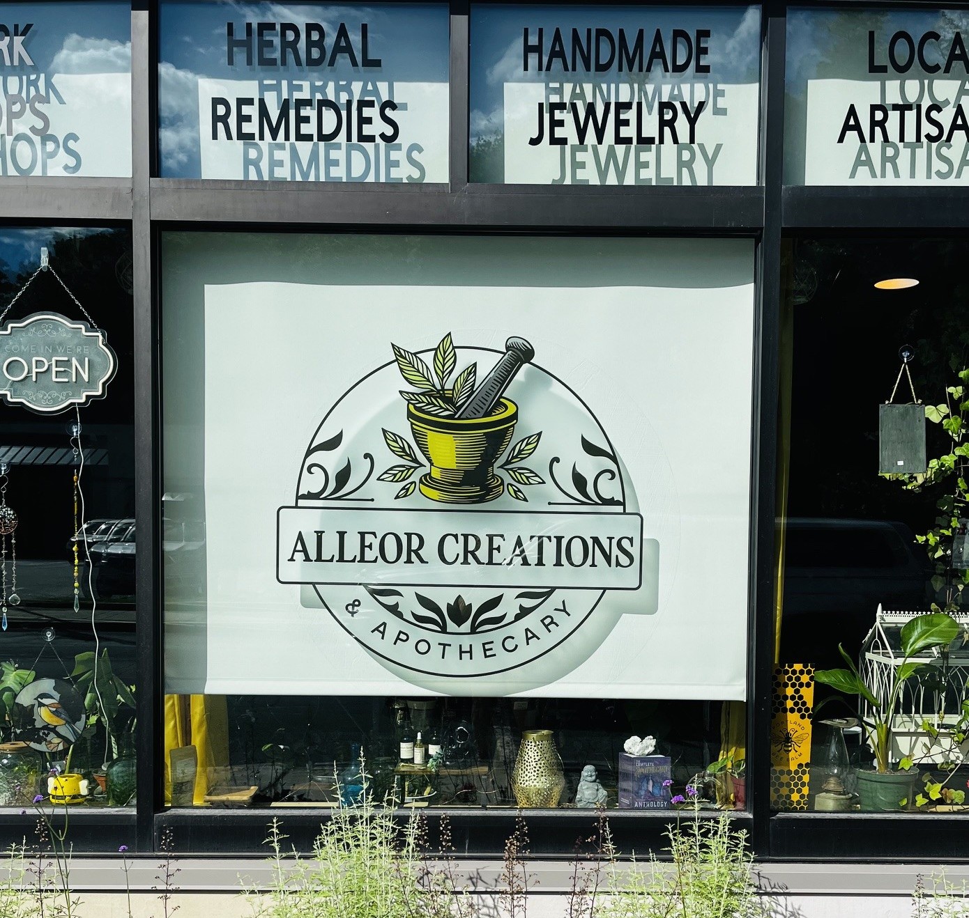 Storefront window display for Alleor Creations Apothecary featuring a circular logo with a mortar and pestle, leaves, and the text 'ALLEOR CREATIONS' and 'APOTHECARY'