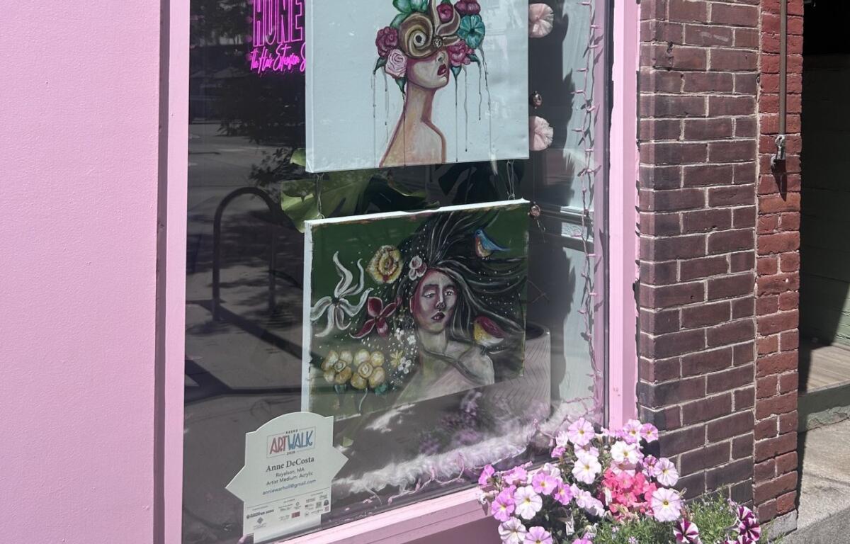 Pink storefront window displaying two hanging art prints: a woman with a flower crown and a surreal portrait, with pink flowers outside the display.