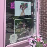 Pink storefront window displaying two hanging art prints: a woman with a flower crown and a surreal portrait, with pink flowers outside the display.