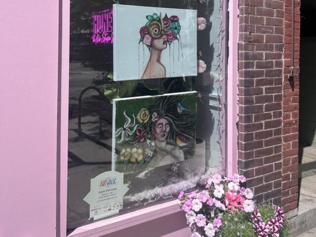 Pink storefront window displaying two hanging art prints: a woman with a flower crown and a surreal portrait, with pink flowers outside the display.