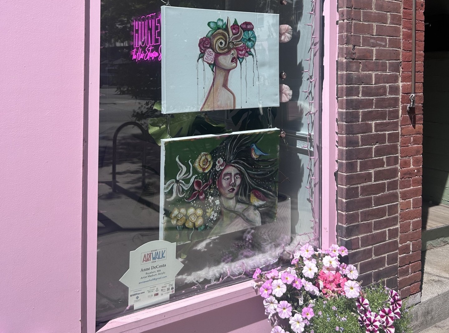 Pink storefront window displaying two hanging art prints: a woman with a flower crown and a surreal portrait, with pink flowers outside the display.