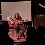 A guitarist with long hair sits on a chair on a dimly lit stage, playing a wooden acoustic guitar in front of a microphone and a blank projection screen, while silhouettes of audience members watch.