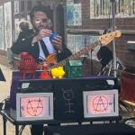 Street musician in sunglasses playing a bass guitar on a station platform, with a prop setup and occult-symbol decorations on the front panel.