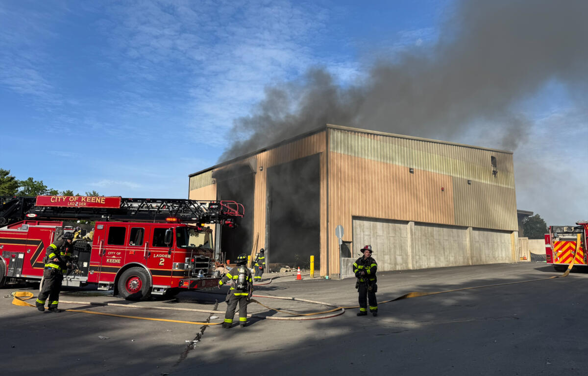 Firefighters battle a warehouse fire as thick smoke billows from the building; a red fire engine is on scene with hoses laid out on the pavement.