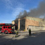 Firefighters battle a warehouse fire as thick smoke billows from the building; a red fire engine is on scene with hoses laid out on the pavement.