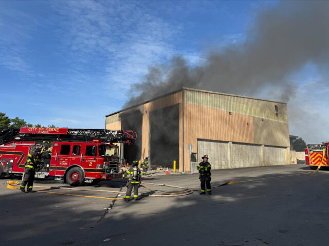Firefighters battle a warehouse fire as thick smoke billows from the building; a red fire engine is on scene with hoses laid out on the pavement.