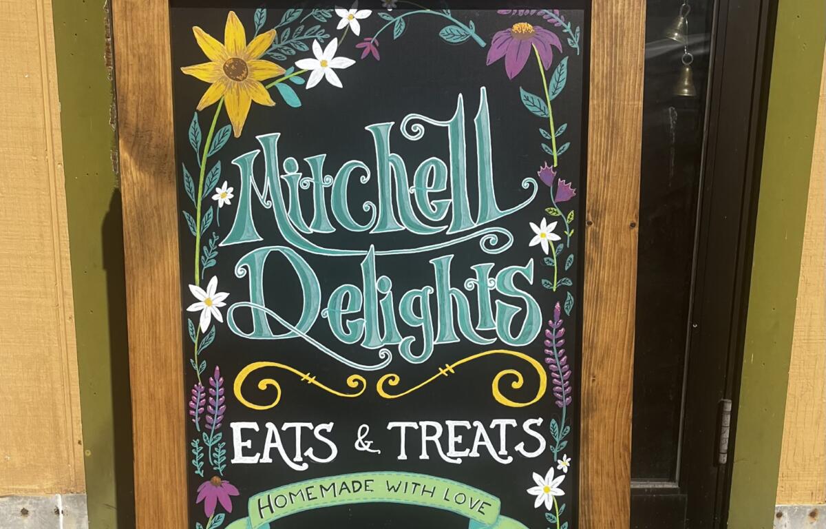 Chalkboard sign on a wooden stand advertising Mitchell Delights: Eats & Treats, homemade with love, with floral border.