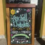 Chalkboard sign on a wooden stand advertising Mitchell Delights: Eats & Treats, homemade with love, with floral border.
