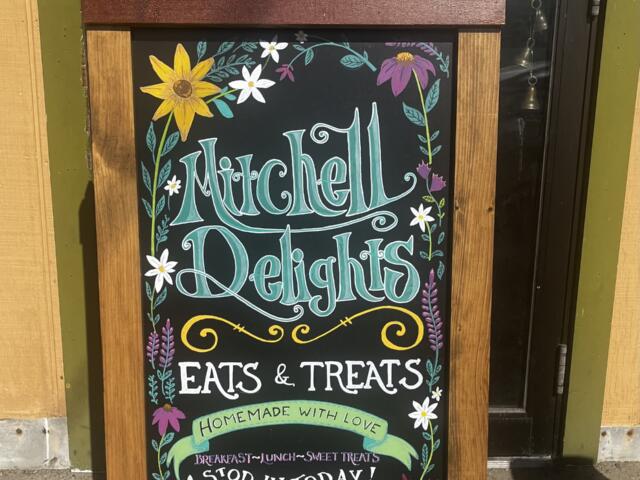 Chalkboard sign on a wooden stand advertising Mitchell Delights: Eats & Treats, homemade with love, with floral border.