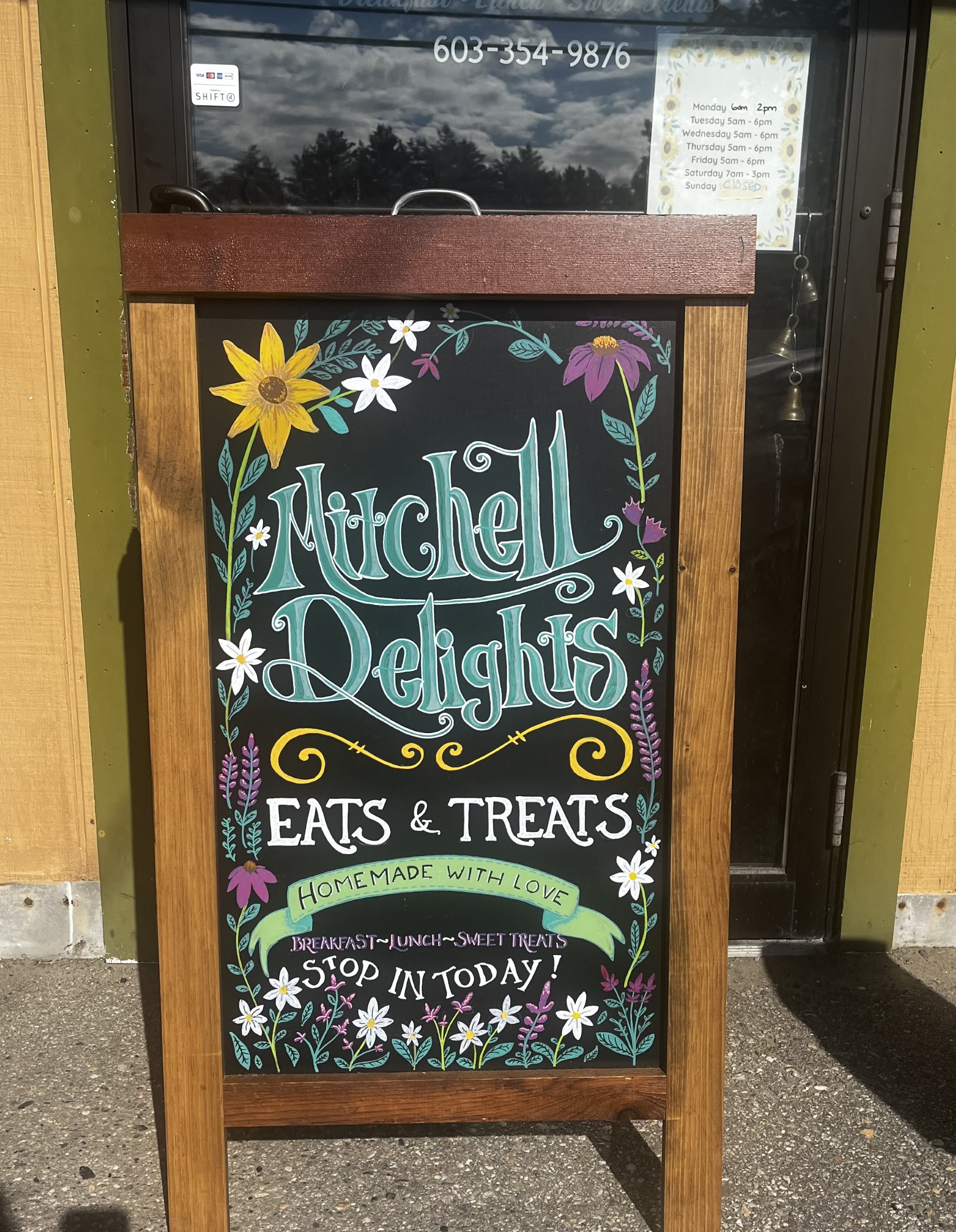 Chalkboard sign on a wooden stand advertising Mitchell Delights: Eats & Treats, homemade with love, with floral border.