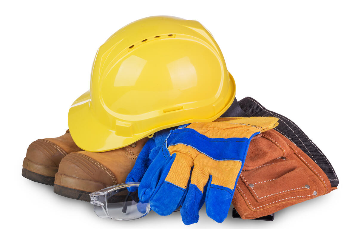 Yellow hard hat atop brown work boots with blue and yellow gloves, safety glasses, and a brown tool pouch stacked together.
