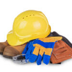 Yellow hard hat atop brown work boots with blue and yellow gloves, safety glasses, and a brown tool pouch stacked together.