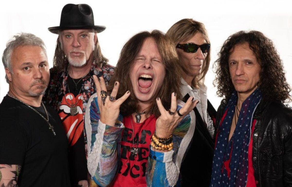 Five rock musicians pose together in a studio, with the central singer shouting and gesturing dramatically, colorful clothing and bracelets visible.