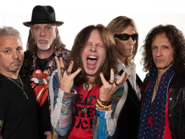 Five rock musicians pose together in a studio, with the central singer shouting and gesturing dramatically, colorful clothing and bracelets visible.