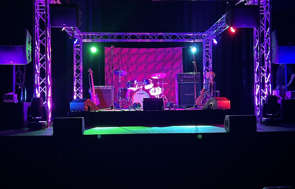 Stage setup for a live performance with purple lighting, drum kit and guitars, and large truss framework above; empty audience in foreground.