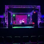 Stage setup for a live performance with purple lighting, drum kit and guitars, and large truss framework above; empty audience in foreground.