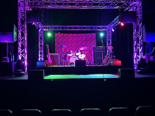 Stage setup for a live performance with purple lighting, drum kit and guitars, and large truss framework above; empty audience in foreground.
