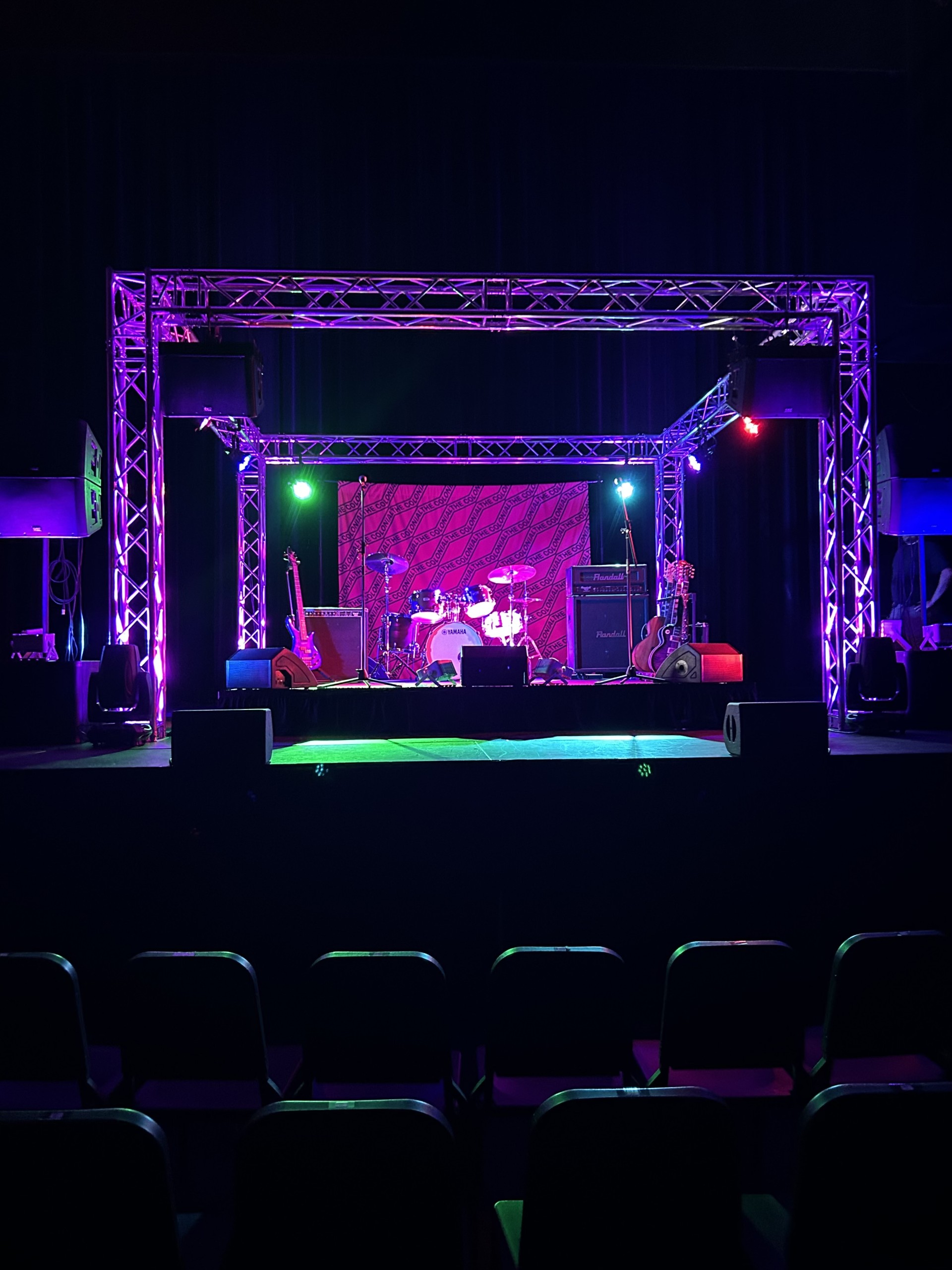 Stage setup for a live performance with purple lighting, drum kit and guitars, and large truss framework above; empty audience in foreground.